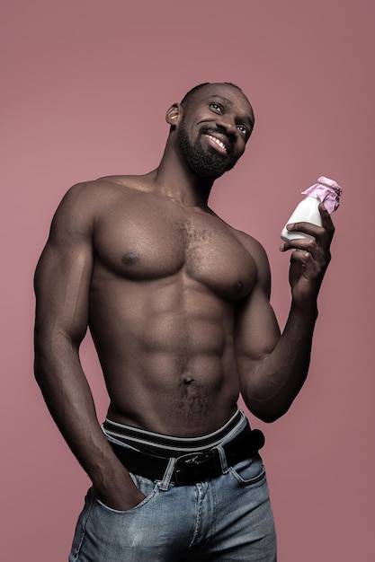 Jeune homme africain en bonne santé tenant une petite bouteille de lait sur un fond de studio rose. Bouchent le portrait dans le style minimaliste d'un jeune homme afro heureux nu