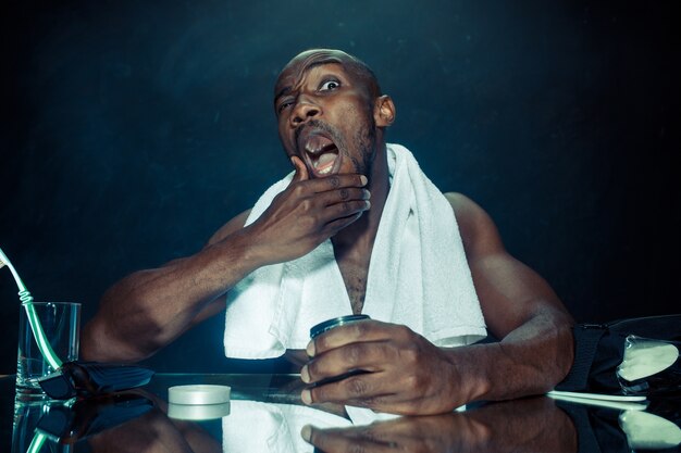 Le jeune homme africain dans la chambre assis devant le miroir après s'être gratté la barbe à la maison. Concept d'émotions humaines. concepts de crème après-rasage