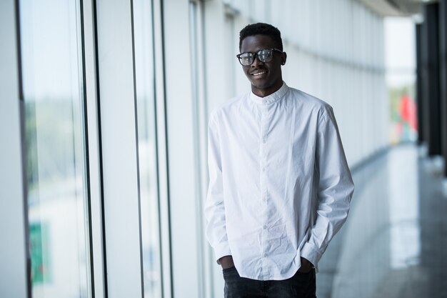 Jeune homme afro-américain à la caméra avec un regard sérieux posant à l'intérieur, debout isolé par une fenêtre blanche