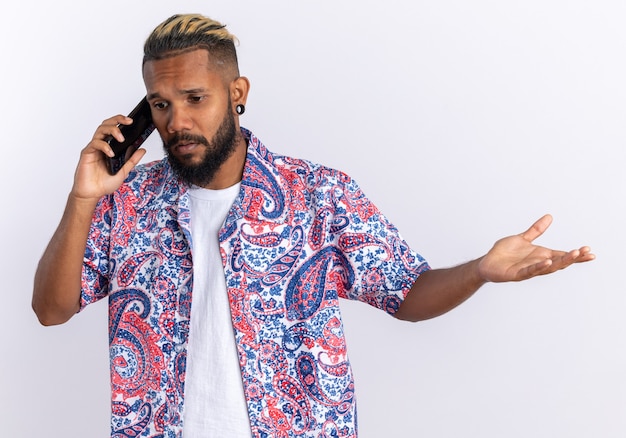 Jeune homme afro-américain en chemise colorée à la confusion tout en parlant au téléphone portable debout sur fond blanc