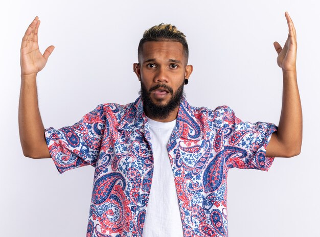 Jeune homme afro-américain en chemise colorée regardant la caméra confuse et mécontente levant les bras dans l'indignation debout sur fond blanc