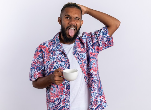 Jeune homme afro-américain en chemise colorée tenant une tasse heureuse et joyeuse qui sort la langue debout sur fond blanc