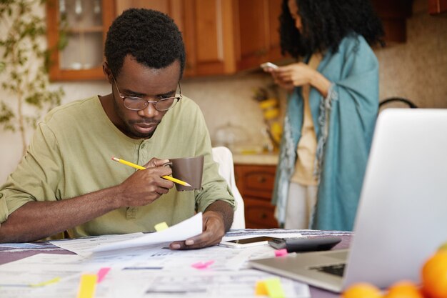 Jeune homme afro-américain dans des verres à boire du café, occupé à travailler sur les finances