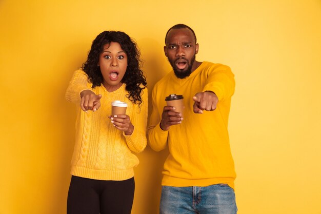 Jeune homme afro-américain émotionnel et femme dans des vêtements décontractés lumineux sur fond jaune. Beau couple. Concept d'émotions humaines, expession faciale, relations, publicité. Boire du café et pointer.