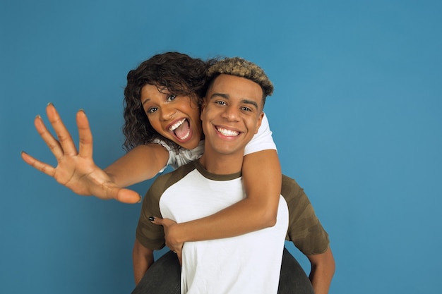 Jeune homme afro-américain émotionnel et femme en vêtements décontractés blancs posant sur fond bleu. Beau couple. Concept d'émotions humaines, expession faciale, relations, publicité. Étreindre, rire.