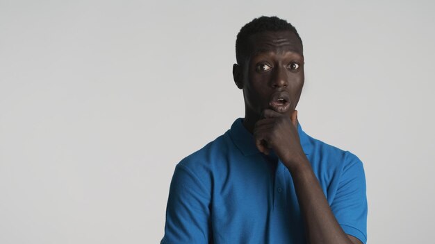 Jeune homme afro-américain étonné se frottant pensivement le menton l'air surpris à huis clos isolé sur fond blanc Wow face à l'expression