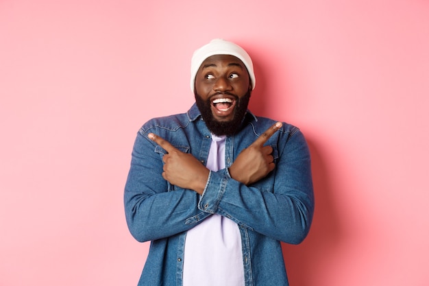 Jeune homme afro-américain excité pointant sur le côté, montrant deux choix et prenant une décision, souriant heureux, debout sur fond rose