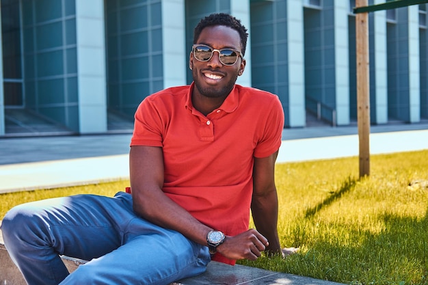 Photo gratuite jeune homme afro-américain heureux en lunettes de soleil est assis sur l'herbe près de gratte-ciel.