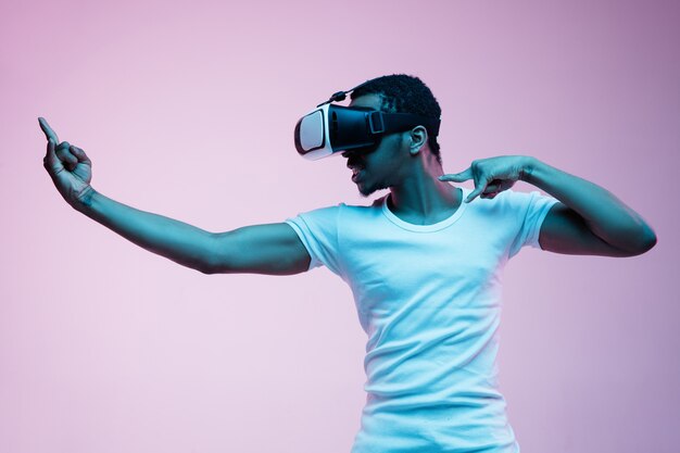 Jeune homme afro-américain jouant dans des lunettes VR en néon sur fond dégradé. Portrait masculin