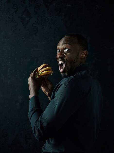 Photo gratuite le jeune homme afro-américain de manger un hamburger et à la recherche de suite sur le studio noir