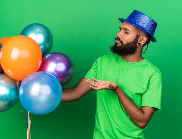 Un jeune homme afro-américain mécontent portant un chapeau de fête tenant et pointant des ballons isolés sur un mur vert