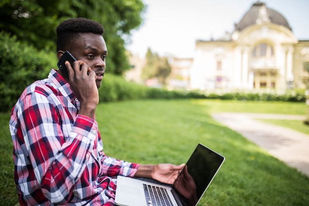 Jeune homme afro-américain parler au téléphone alors qu'il était assis sur l'herbe verte avec un ordinateur portable sur la rue de la ville