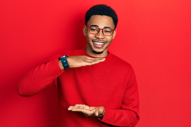 Jeune homme afro-américain portant des vêtements décontractés et des lunettes gesticulant avec les mains montrant le symbole de mesure de signe de grande et grande taille souriant en regardant le concept de mesure de la caméra