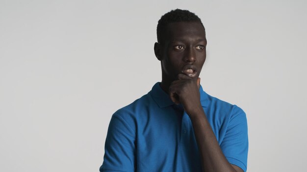 Jeune homme afro-américain réfléchi frottant le menton en pensant à la caméra sur fond blanc Profonde dans l'expression de la pensée