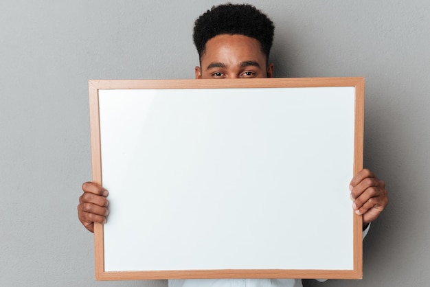 Photo gratuite jeune homme afro-américain se cachant derrière une planche vierge