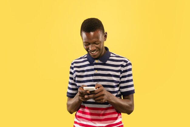 Jeune homme afro-américain avec smartphone isolé sur fond de studio jaune, expression faciale. Beau portrait d'homme demi-longueur. Concept d'émotions humaines, expression faciale.