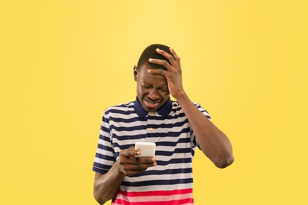 Jeune homme afro-américain avec smartphone isolé sur fond de studio jaune, expression faciale. Beau portrait d'homme demi-longueur. Concept d'émotions humaines, expression faciale.