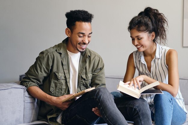 Jeune homme afro-américain souriant et jolie femme asiatique lisant joyeusement un livre ensemble dans un espace de coworking moderne