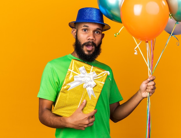 Jeune homme afro-américain surpris portant un chapeau de fête tenant des ballons avec une boîte-cadeau isolée sur un mur orange