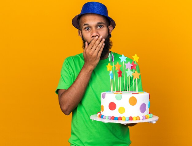 Jeune homme afro-américain surpris portant un chapeau de fête tenant un visage couvert de gâteau avec une main isolée sur un mur orange