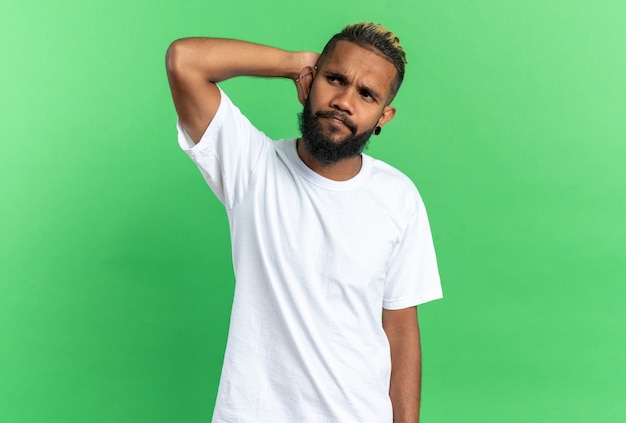 Jeune homme afro-américain en t-shirt blanc à côté perplexe avec la main sur la tête debout sur fond vert