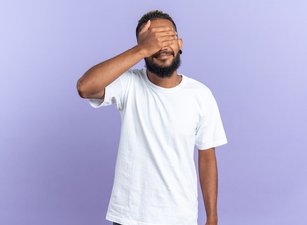 Jeune homme afro-américain en t-shirt blanc fermant les yeux avec la main debout sur fond bleu