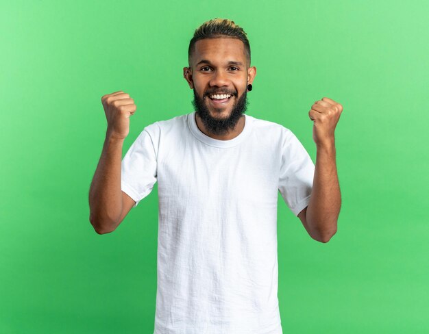 Jeune homme afro-américain en t-shirt blanc heureux et excité serrant les poings se réjouissant de son succès debout sur fond vert