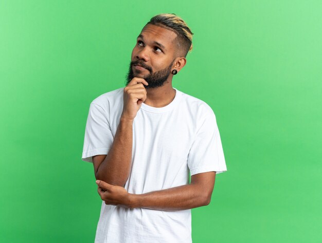 Jeune homme afro-américain en t-shirt blanc levant perplexe avec la main sur son menton debout sur fond vert