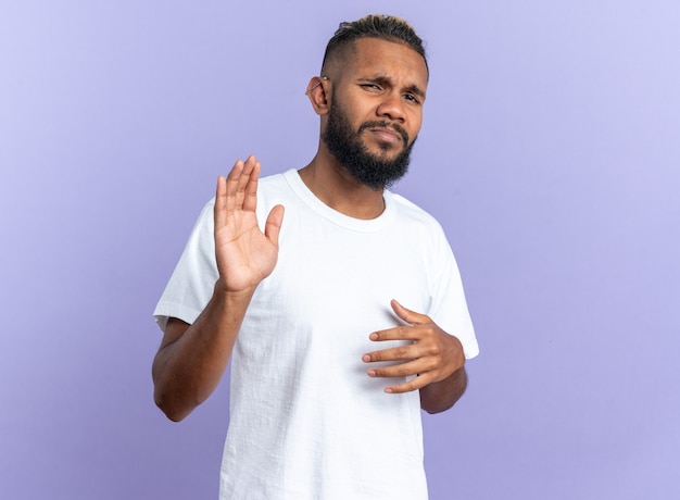 Jeune homme afro-américain en t-shirt blanc regardant la caméra confus et mécontent de faire un geste d'arrêt avec les mains debout sur fond bleu