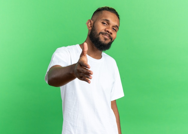 Jeune homme afro-américain en t-shirt blanc regardant la caméra souriant amical offrant des salutations à la main debout sur fond vert