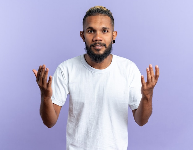 Jeune homme afro-américain en t-shirt blanc regardant la caméra avec un visage heureux levant les bras debout sur fond bleu