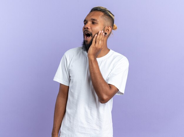 Jeune homme afro-américain en t-shirt blanc regardant de côté étonné et surpris debout sur bleu