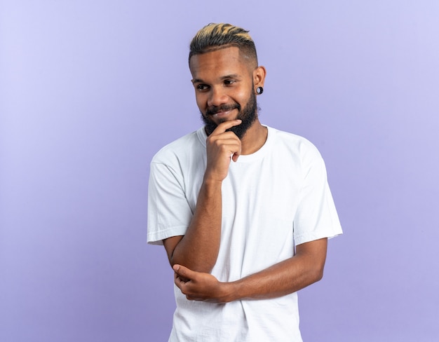 Jeune homme afro-américain en t-shirt blanc regardant de côté avec la main sur le menton souriant confiant debout sur fond bleu