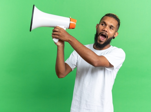 Photo gratuite jeune homme afro-américain en t-shirt blanc tenant un mégaphone criant émotionnellement et heureux debout sur fond vert
