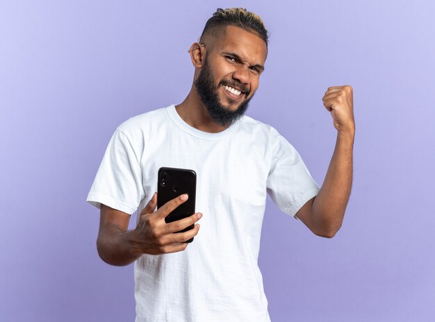 Jeune homme afro-américain en t-shirt blanc tenant un smartphone serrant le poing heureux