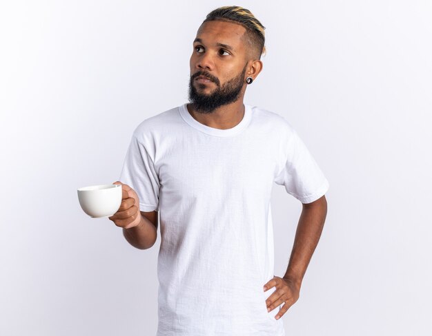 Jeune homme afro-américain en t-shirt blanc tenant une tasse de thé regardant de côté avec un visage sérieux debout sur blanc