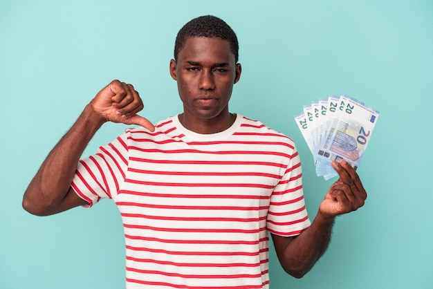 Jeune homme afro-américain tenant un billet de banque isolé sur fond bleu montrant un geste d'aversion, les pouces vers le bas. notion de désaccord.
