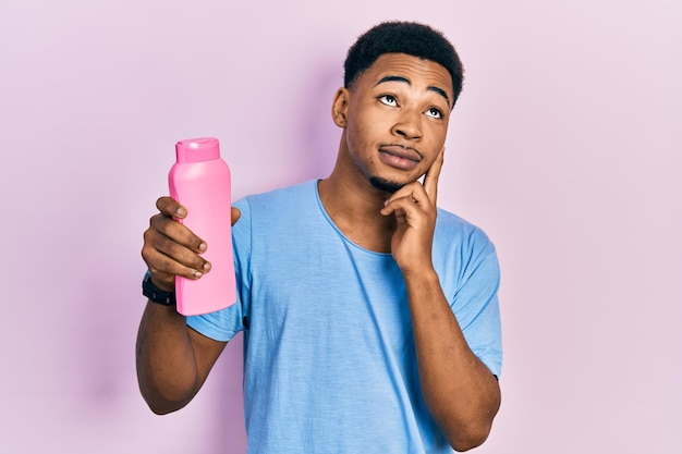 Jeune homme afro-américain tenant une bouteille de shampoing visage sérieux pensant à la question avec la main sur le menton, pensif à l'idée confuse