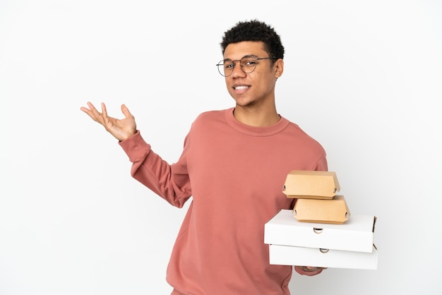 Jeune homme afro-américain tenant un hamburger et des pizzas isolés sur fond blanc tendant les mains sur le côté pour inviter à venir