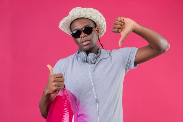 Jeune homme afro-américain voyageur en chapeau d'été avec des écouteurs autour de son cou portant des lunettes de soleil noires tenant un anneau gonflable montrant les pouces de haut en bas sur p