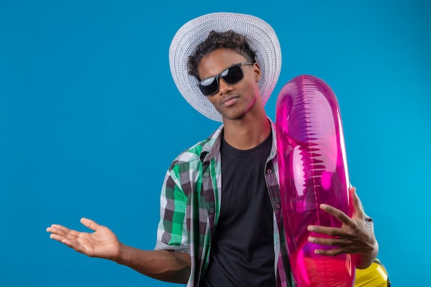 Jeune homme afro-américain voyageur en chapeau d'été portant des lunettes de soleil noires tenant anneau gonflable présentant avec bras de sa main copie espace regardant la caméra avec un sourire confiant o