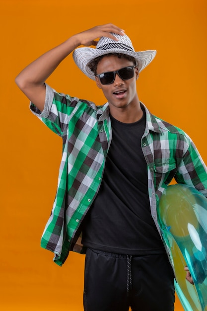 Photo gratuite jeune homme afro-américain voyageur en chapeau d'été portant des lunettes de soleil noires tenant l'anneau gonflable sorti en regardant la caméra en touchant son chapeau debout sur fond orange