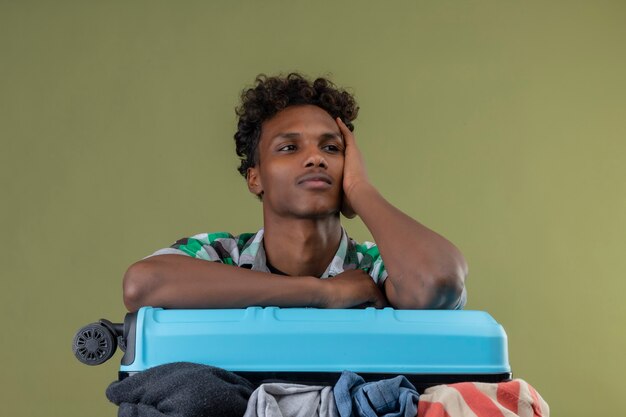Jeune homme afro-américain voyageur debout avec une valise pleine de vêtements à la recherche de côté avec une expression pensive pensée positive sur fond vert