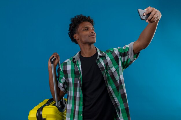 Jeune homme afro-américain voyageur debout avec valise prenant selfie à l'aide de son smartphone debout sur fond bleu