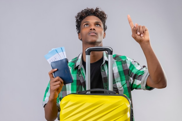 Jeune homme afro-américain voyageur debout avec valise tenant des billets d'avion à la recherche et pointant le doigt vers le haut avec un visage sérieux sur fond blanc