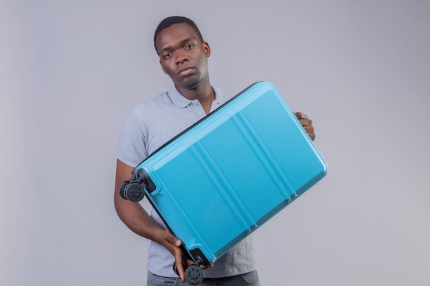Jeune homme afro-américain voyageur en polo gris tenant une valise bleue avec visage malheureux