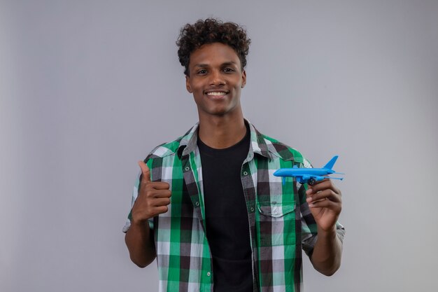 Jeune homme afro-américain voyageur tenant avion jouet souriant positif et heureux montrant les pouces vers le haut debout sur fond blanc