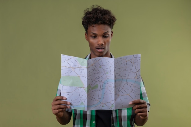 Jeune homme afro-américain voyageur tenant une carte en le regardant surpris debout sur fond vert