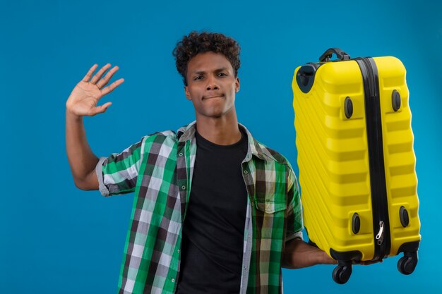 Jeune homme afro-américain voyageur tenant valise en agitant avec la main en regardant les caméras avec une expression sceptique sur le visage debout sur fond bleu