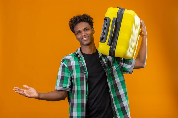 Jeune homme afro-américain voyageur tenant valise regardant la caméra souriant positif et heureux propagation des mains faisant un geste de bienvenue debout sur fond orange